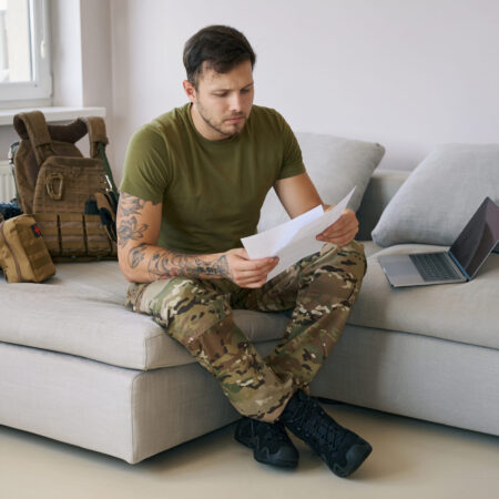 Military serviceman studying documents in hands while sitting on couch next to laptop and army gear